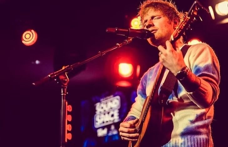 Ed Sheeran performing onstage, playing acoustic guitar and singing into a microphone. He wears a tie-dye sweater, lit by stage lights.