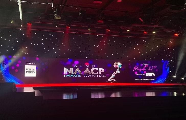 Stage setup for the 53rd NAACP Image Awards. A person stands center stage in front of a large screen, backdrop with lights and event logo.