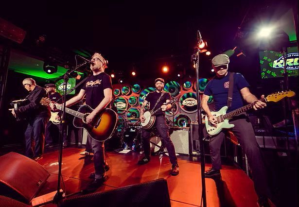A band of five men performing on stage, illuminated by green and orange lights. Two guitarists, a banjo player, a bassist, and an accordion player are shown.
