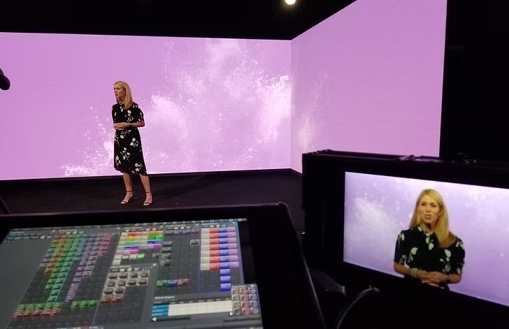 Woman on stage presenting in front of a purple background. A control panel and monitor displaying the presenter are in the foreground.