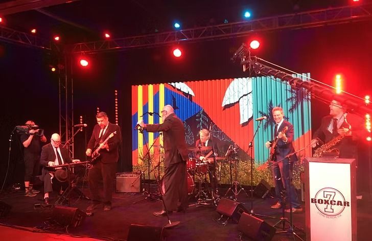 A band performs on stage with colorful background graphics. The band members are dressed in suits and lit with red stage lights.