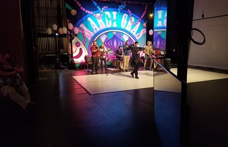 A band performs on a stage decorated for Mardi Gras. A man in a black suit dances center stage.