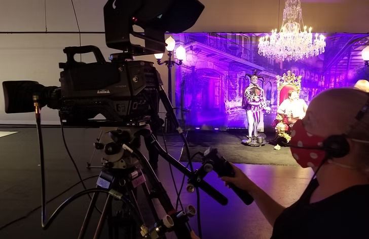 Camera operator filming performers on a stage with purple lighting, a throne, and a chandelier. The operator wears a face mask and headphones.