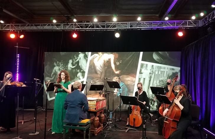 An ensemble performs on stage with a projected backdrop. The musicians are dressed in formal attire and face their instruments. The setting includes stage lights and curtains.