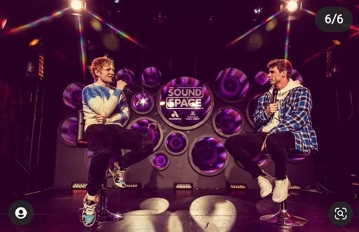 Ed Sheeran and a man in plaid jacket sit on stools, microphones in hand, on stage with purple lighting and