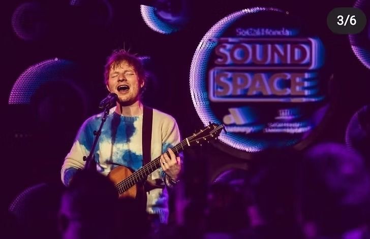 Ed Sheeran performing onstage at Sound Space, playing guitar, wearing a tie-dye sweater, with purple lighting and background.