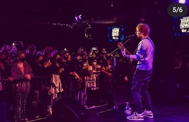 Ed Sheeran performing on stage at a concert, playing guitar. The audience is in the dark, with many wearing masks, holding up phones.
