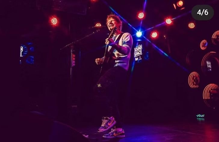 A musician on stage playing guitar, bathed in stage lights. He wears a t-shirt, black pants, and patterned sneakers.