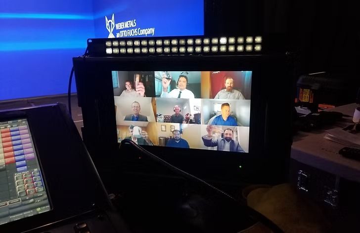 A screen displays a video call with nine people. Blue background, dark setting, and production equipment visible.
