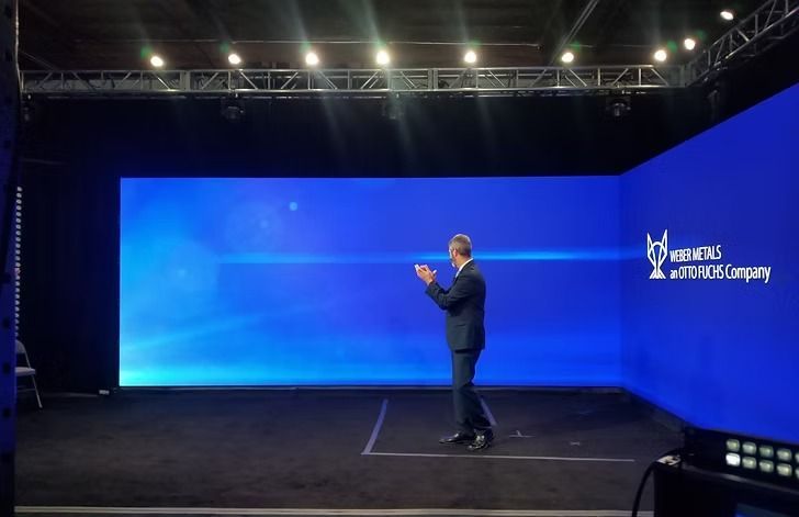 Man in suit clapping on stage with a large blue screen, company logo visible. Dark setting with stage lights.