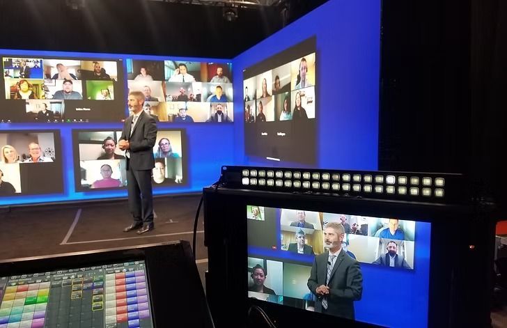 Man in suit giving a presentation on a stage with multiple screens displaying video call participants; a control panel and monitor are in the foreground.