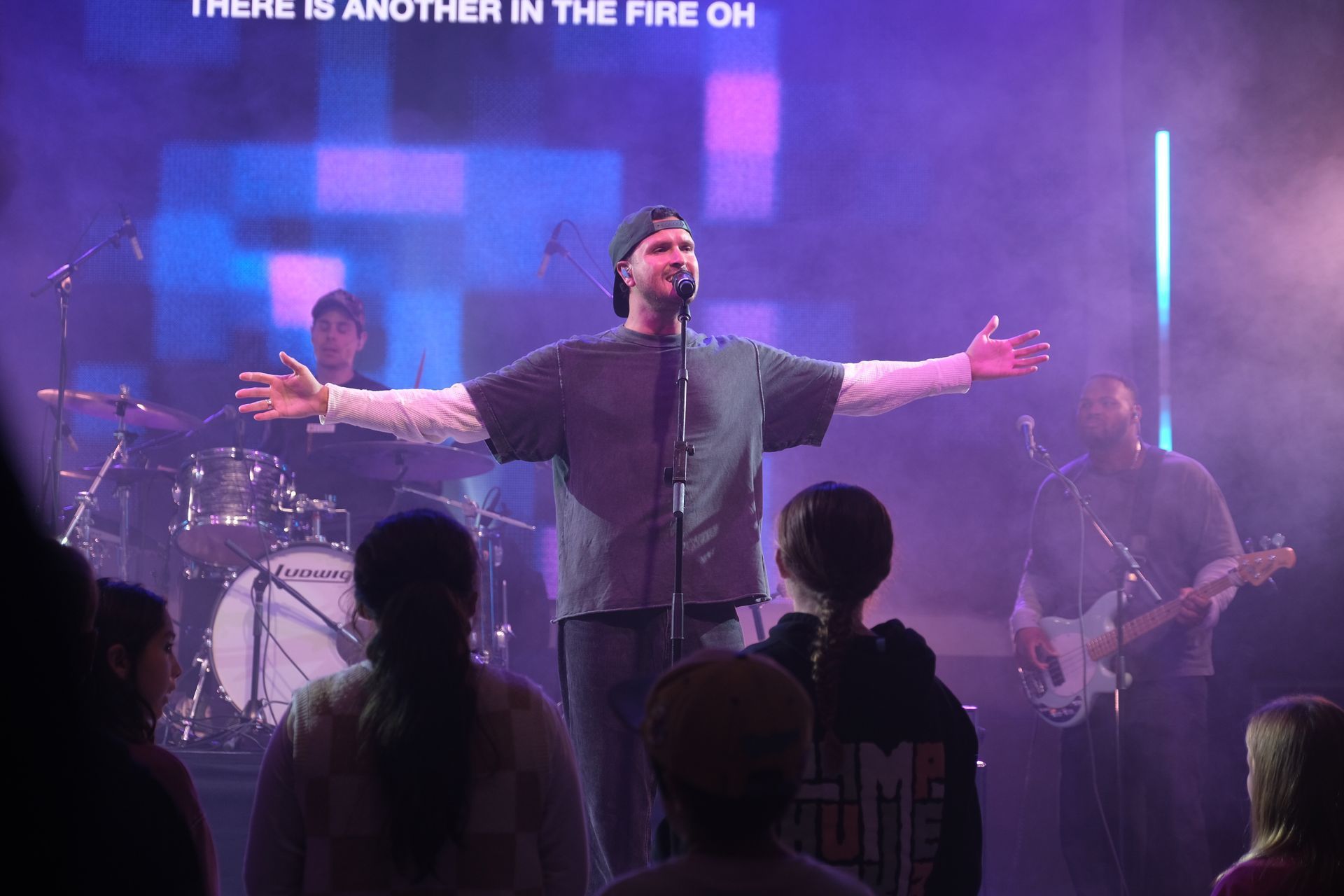 Man singing on stage with arms outstretched, band and audience. Blue and purple stage lighting.
