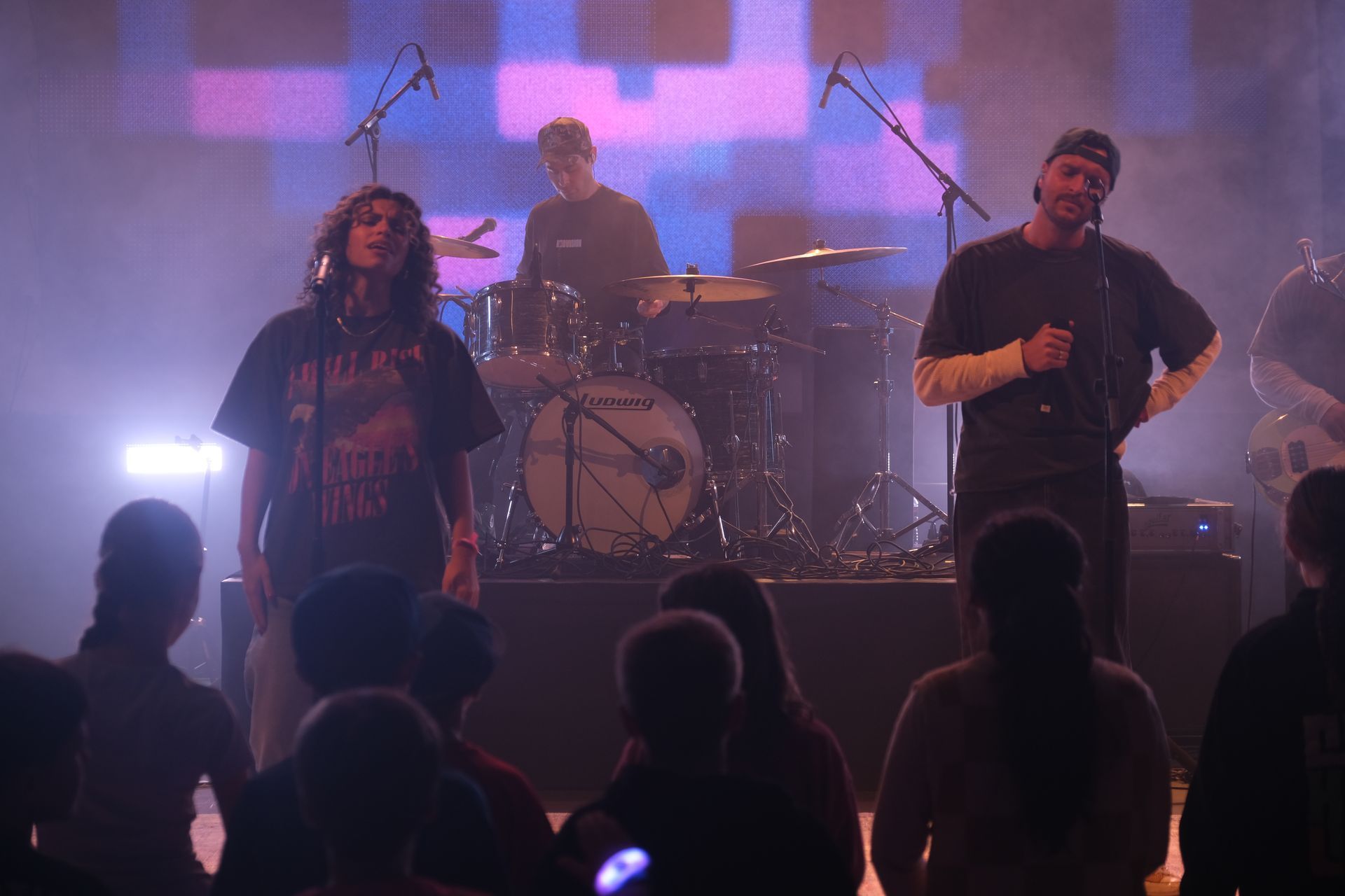 A band performs on stage. Two vocalists and a drummer are visible, with an audience in front.