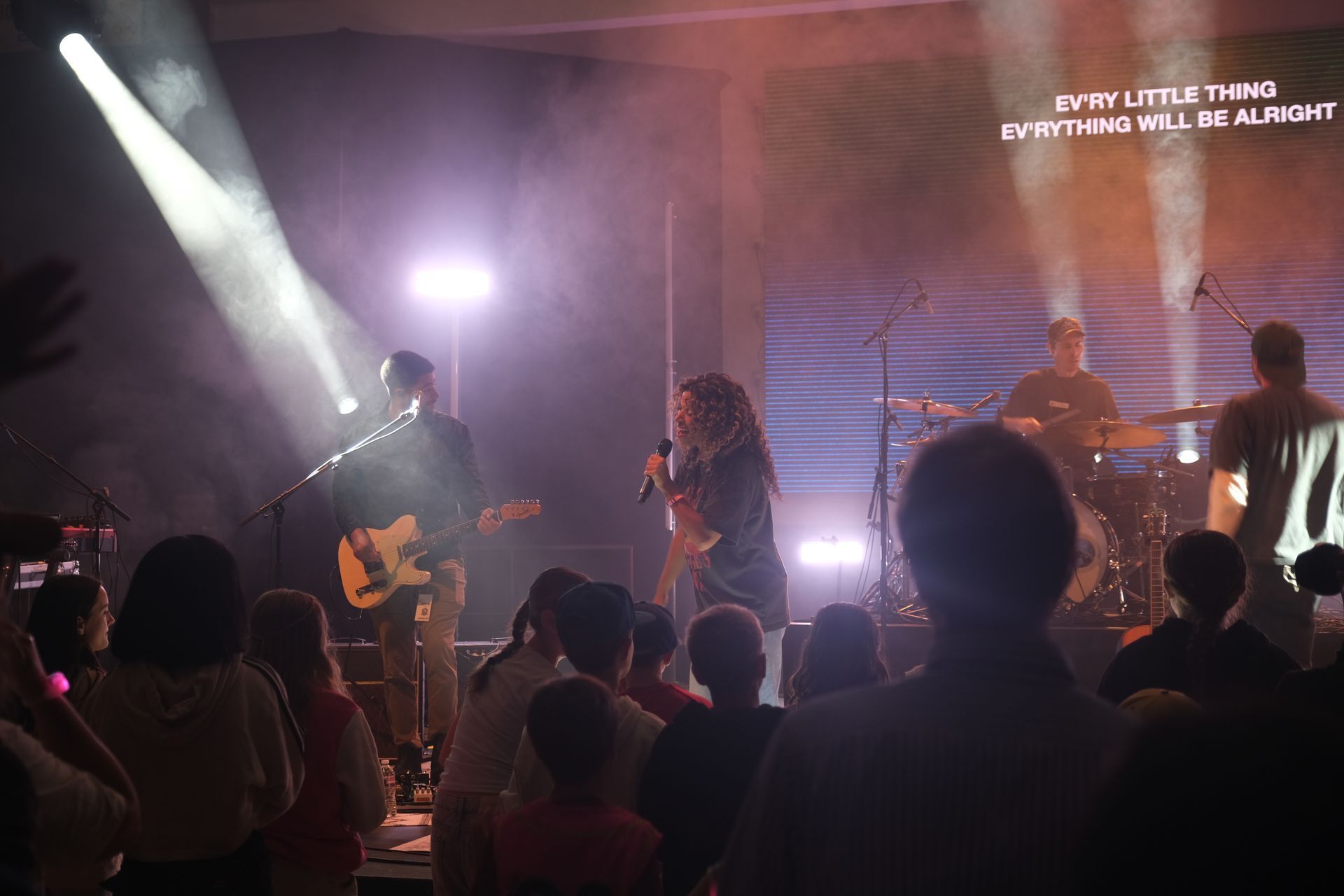 Band performing on stage with audience. Stage is lit, with lyrics projected on screen.