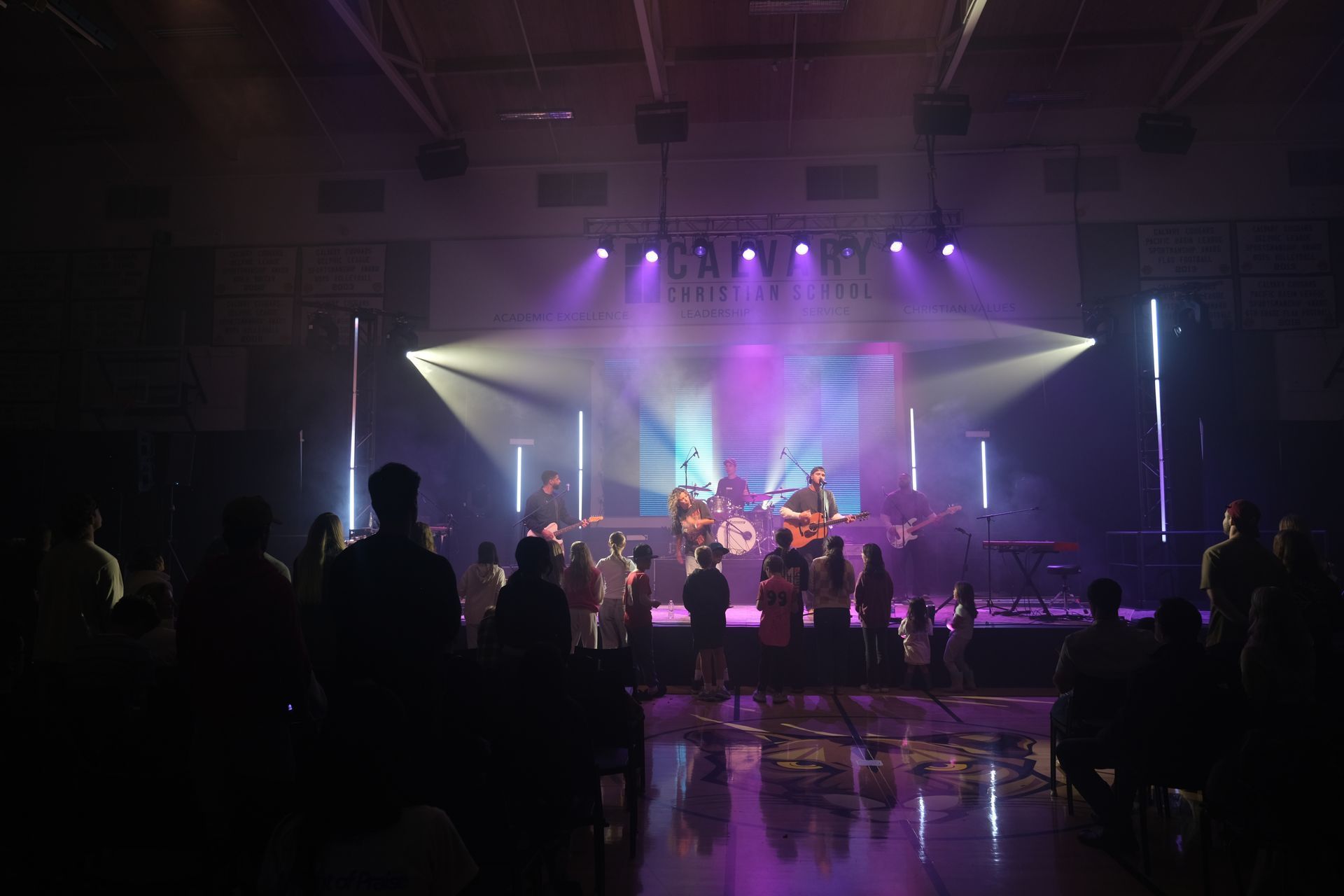 A band performs on stage with purple and white stage lights; audience watches in the dark.