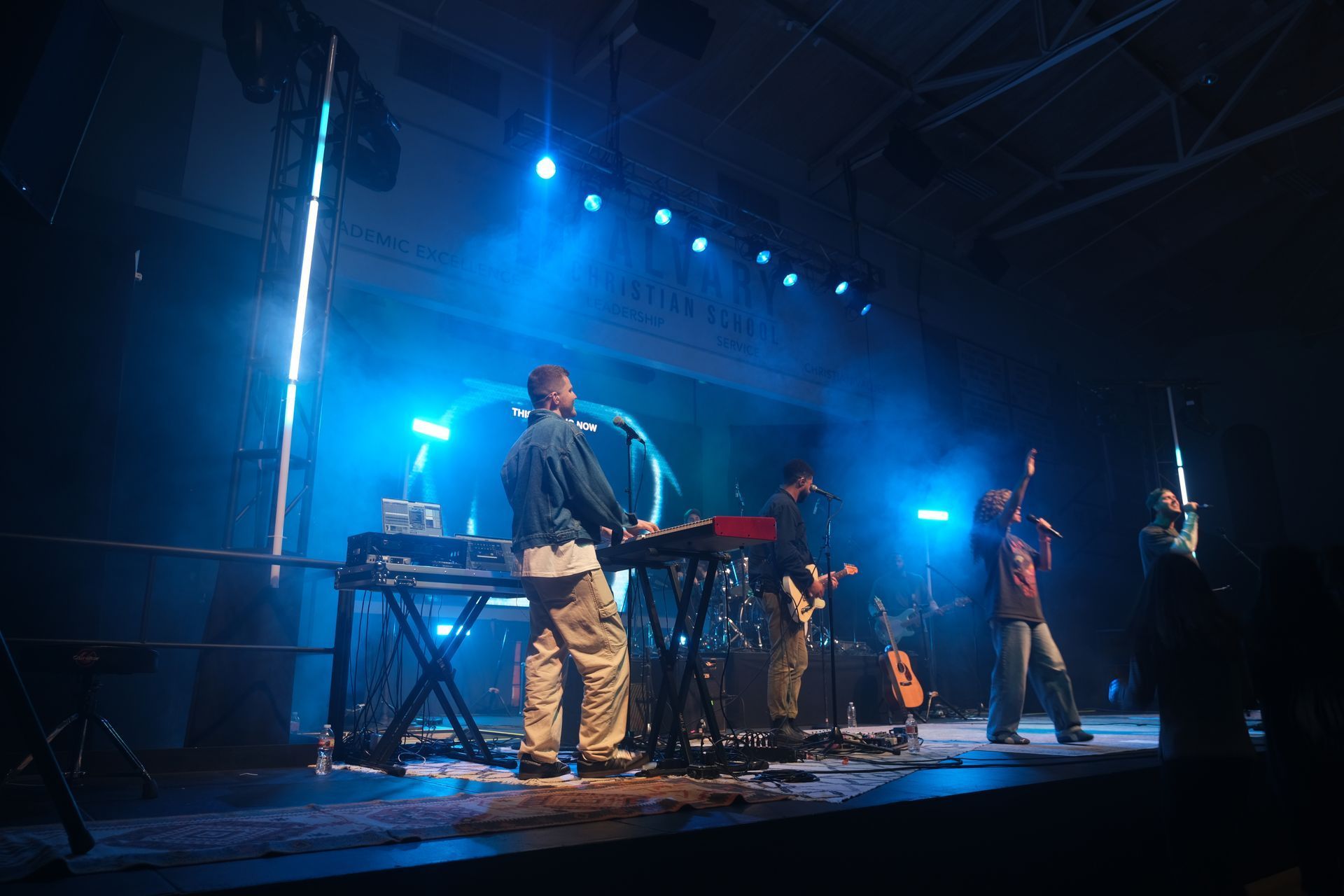 Band performing on stage, bathed in blue lights. Musician at keyboard, singer with microphone, drummer, other band members.