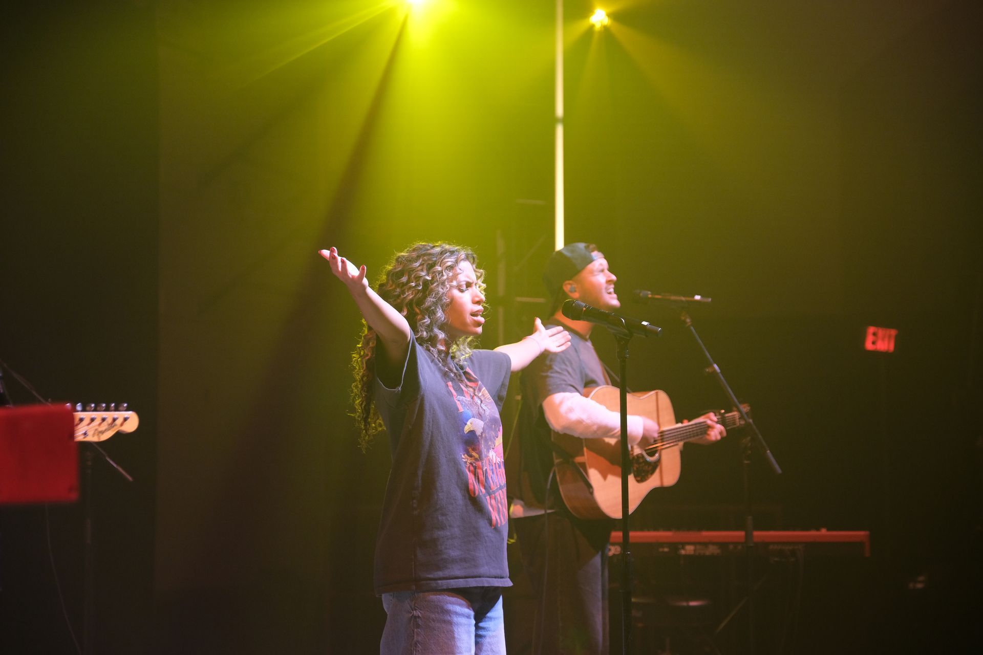Two people on stage performing. Woman with arms raised, man playing guitar, both under yellow spotlights.