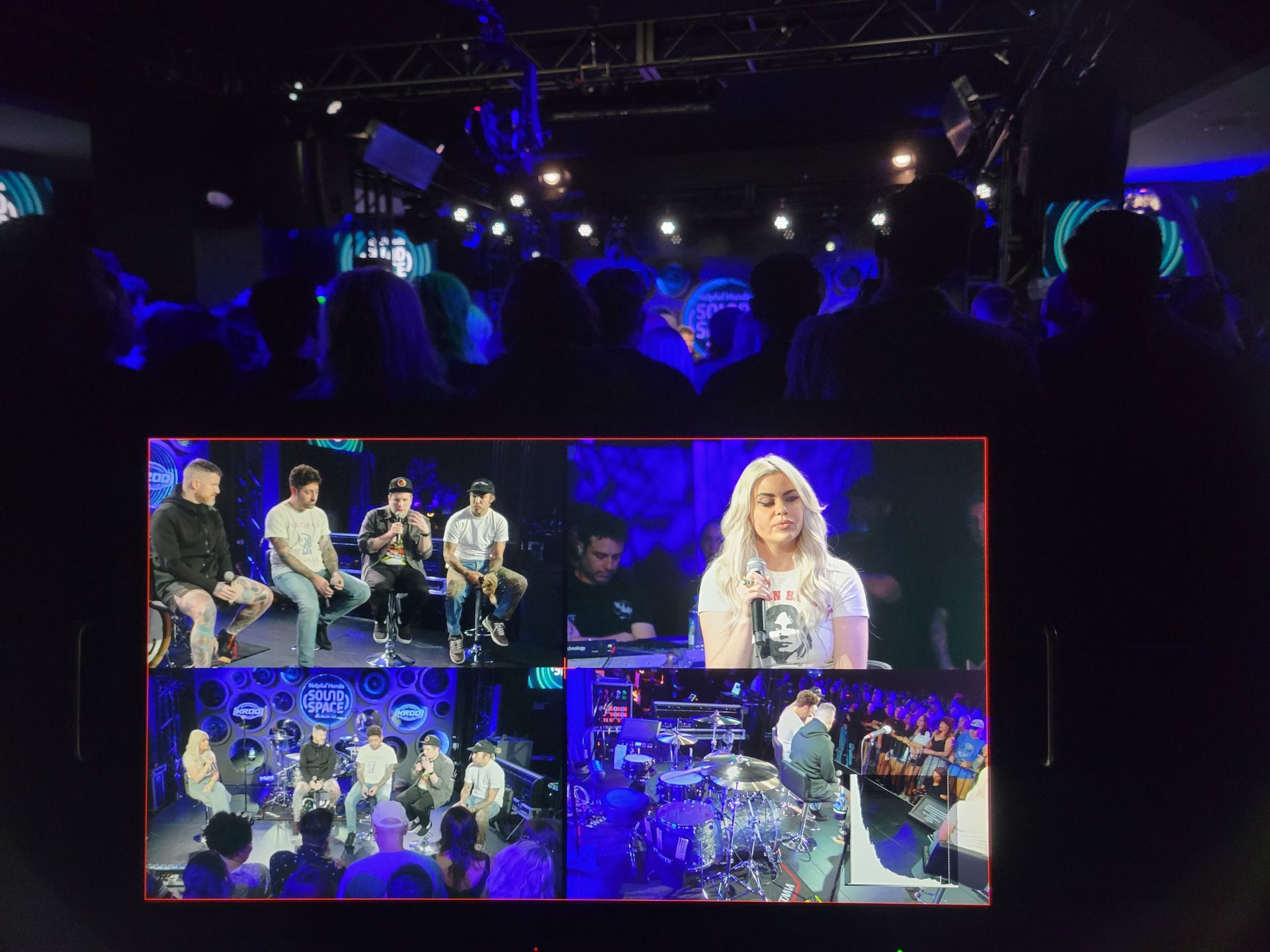 A stage with an audience and a screen showing a live event: panel discussion, interview, and drumming performance.