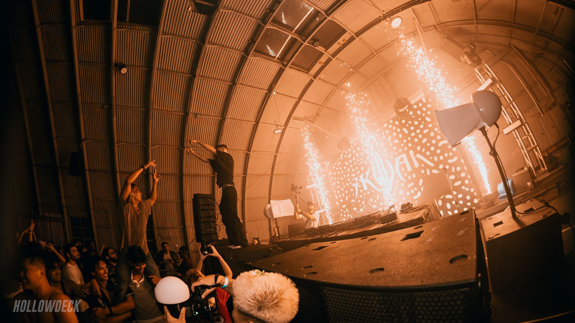 DJ performs on stage with pyrotechnics. Crowd watches in a domed, industrial-looking venue.