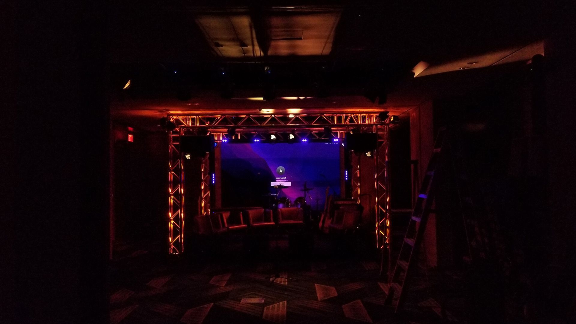 Dark hallway leading to a stage with a large screen and orange and blue stage lights.