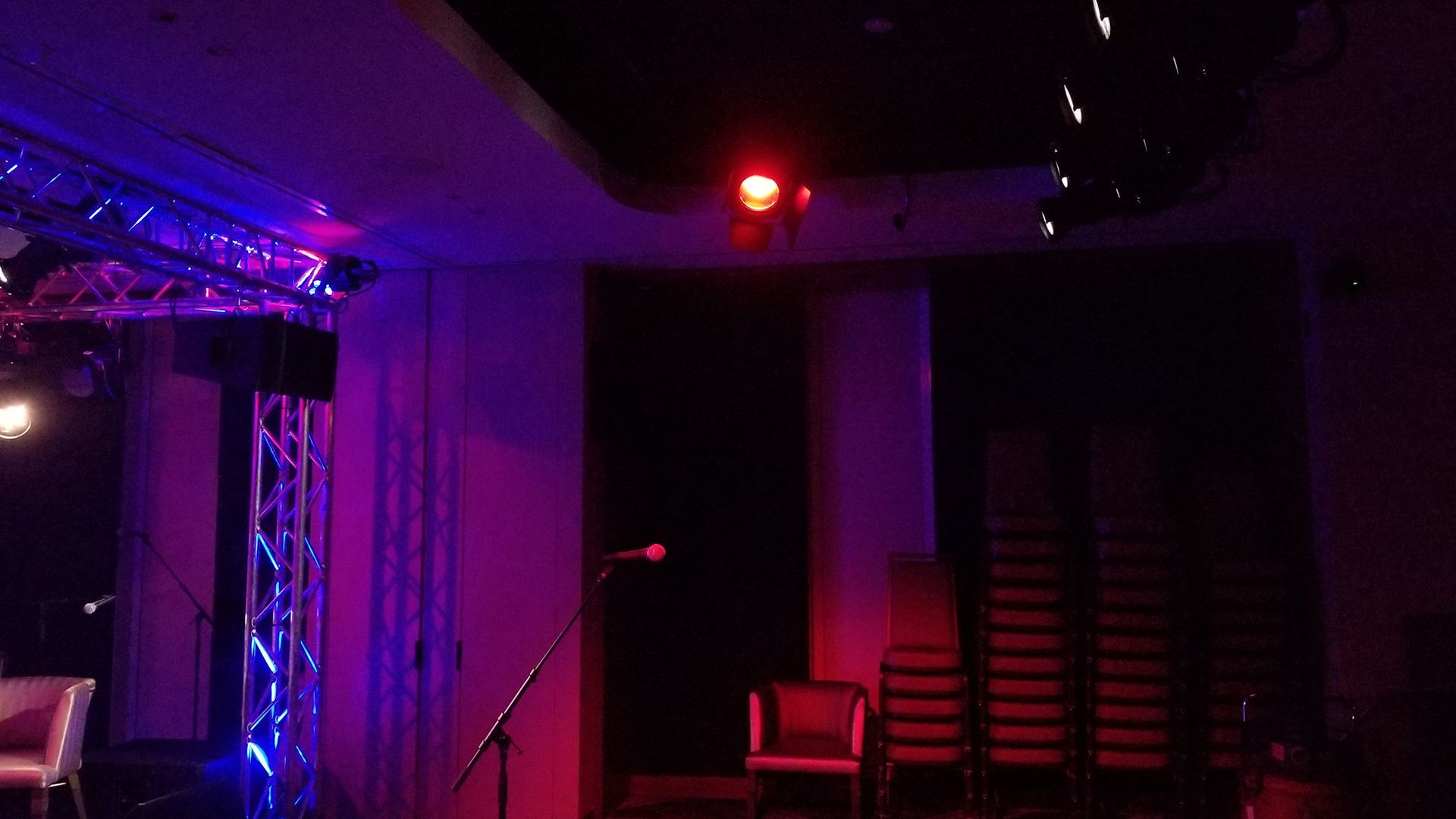 A dimly lit stage with blue and red stage lights, microphone, chairs, and a stack of chairs.
