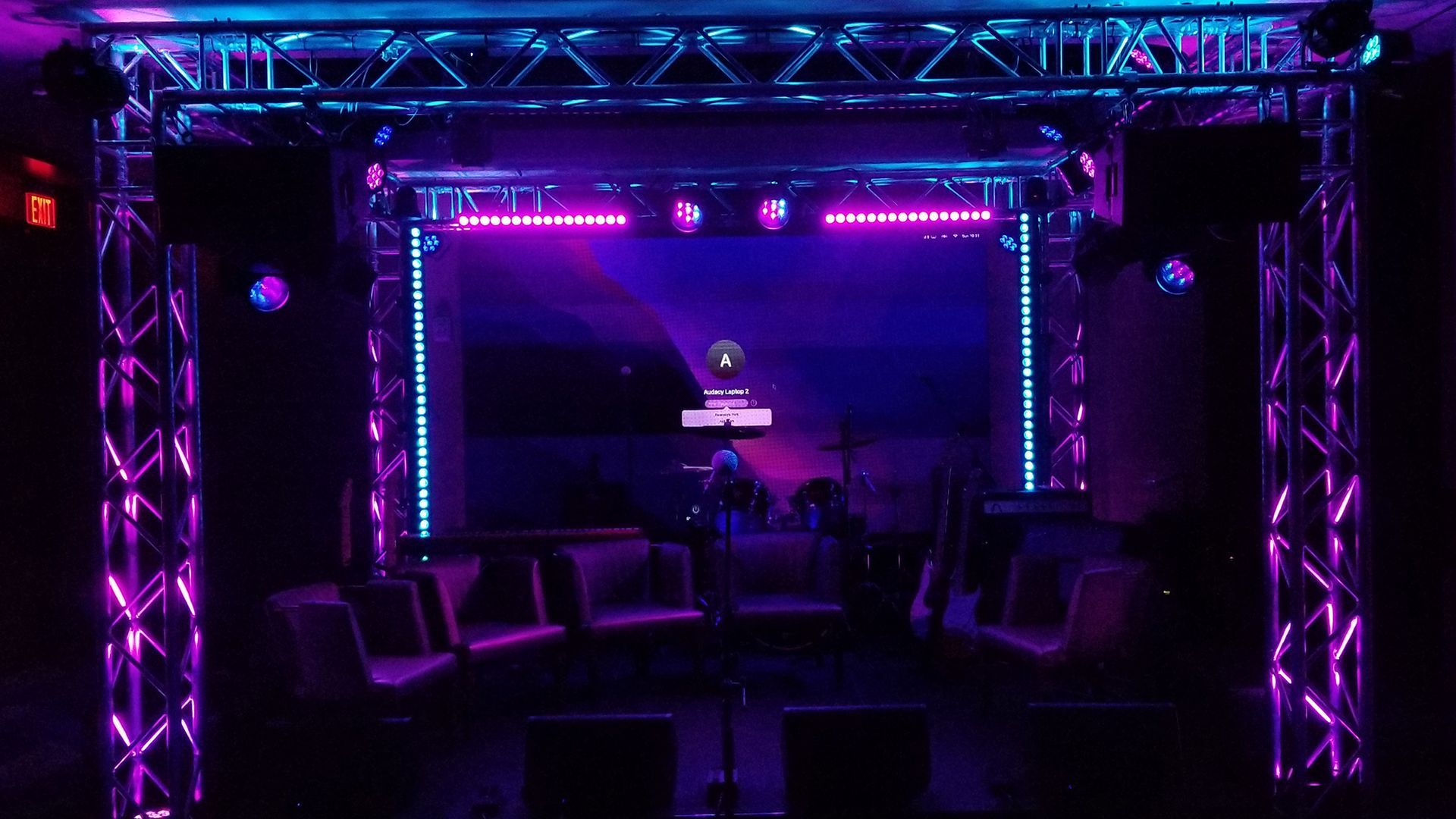 A stage setup with purple and blue lighting, trusses, and sound equipment. Chairs are in the foreground.