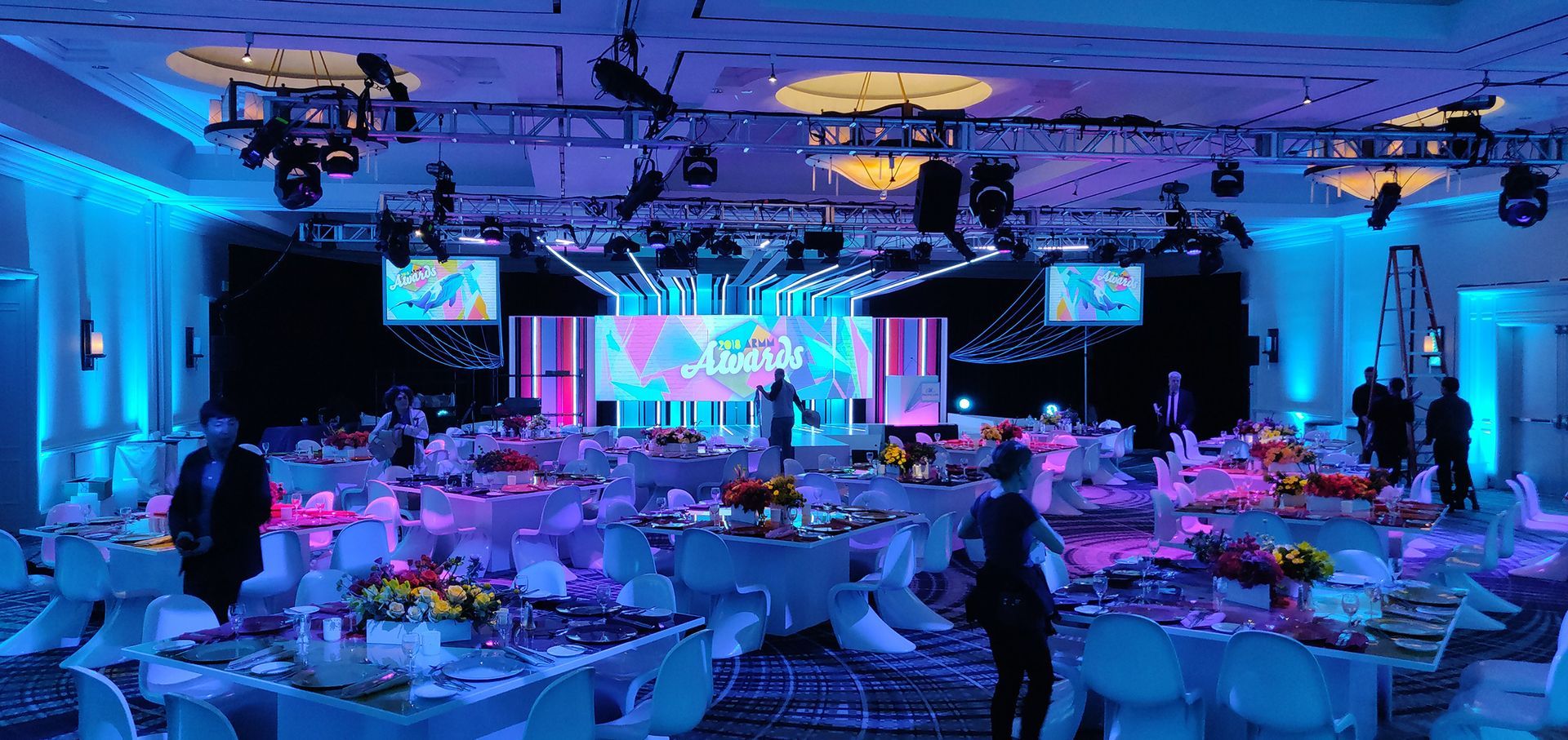 A brightly lit ballroom with tables set for a gala. A person is on a stage in front of a screen.