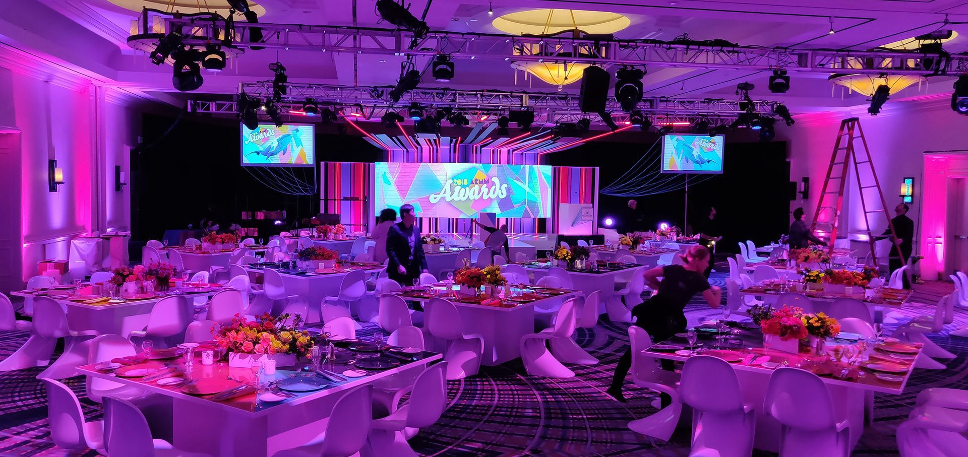 A lavish event hall, tables set for a gala, bathed in purple light, stage at the back.