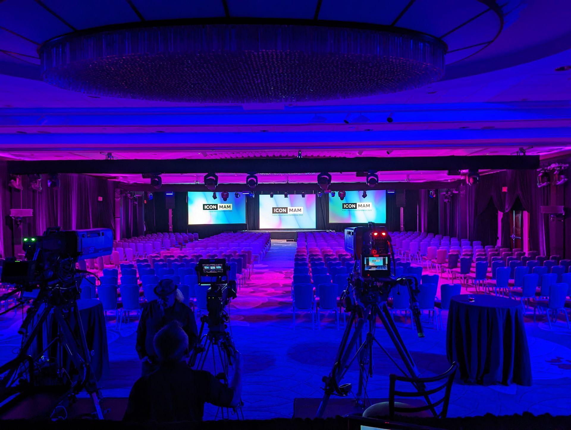 Conference room setup: Three screens, rows of chairs, cameras, blue and purple lighting.