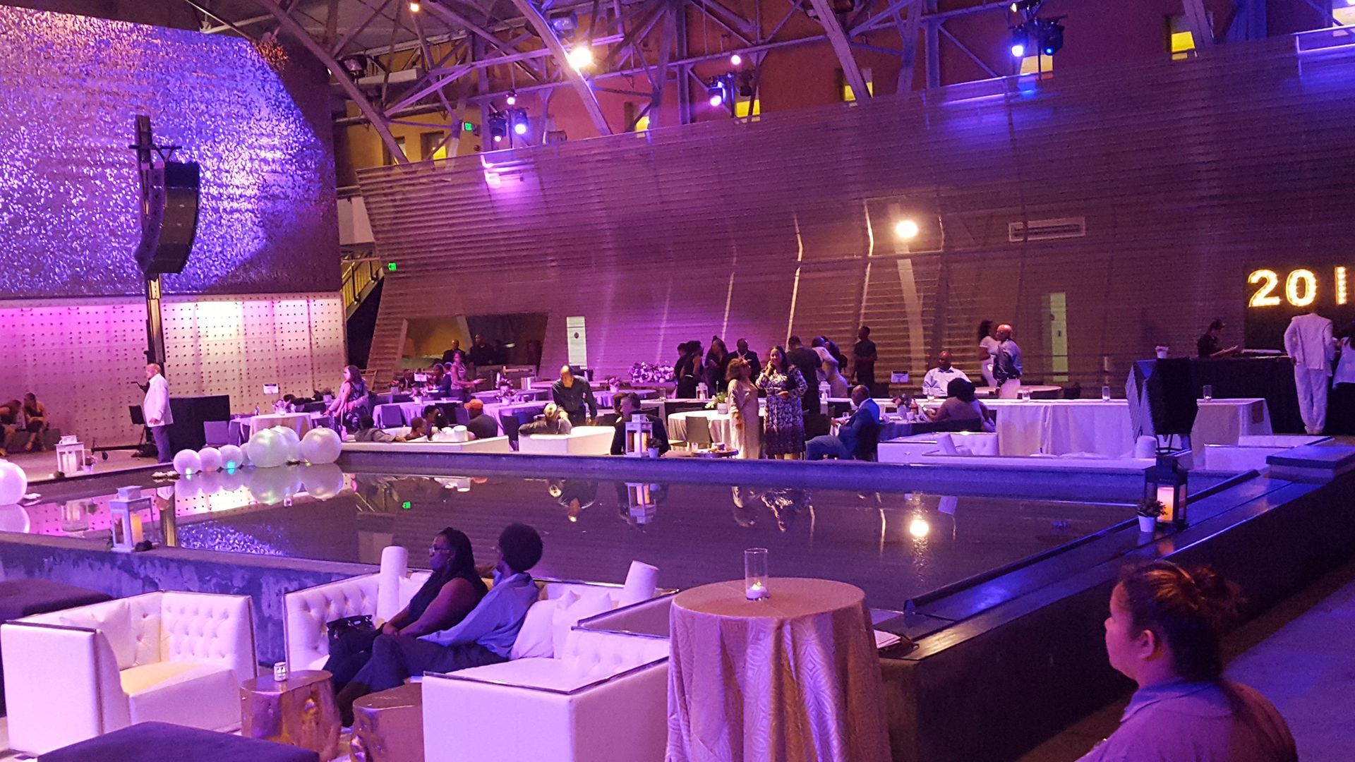 Event venue with seated guests near a reflecting pool. Purple lighting and a stage in the background.