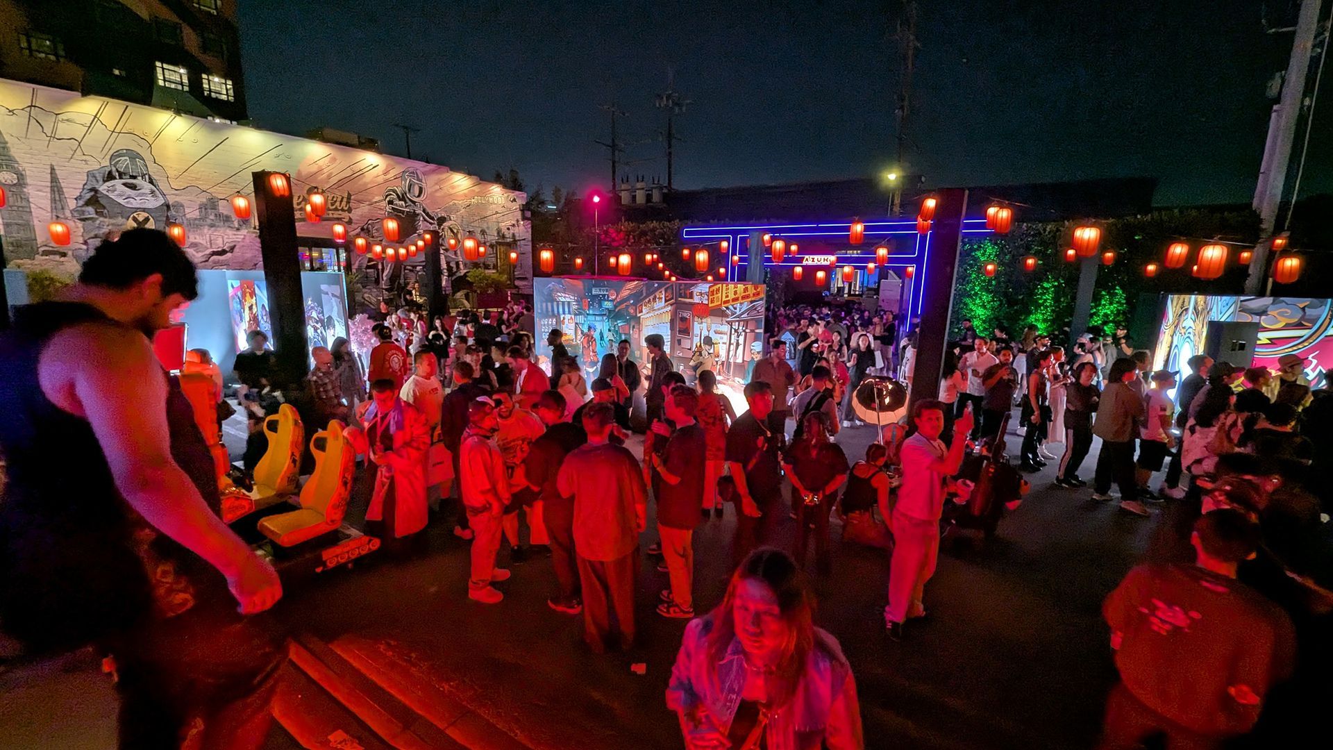 Night scene of a crowded outdoor event with people, lights, and artwork on walls.