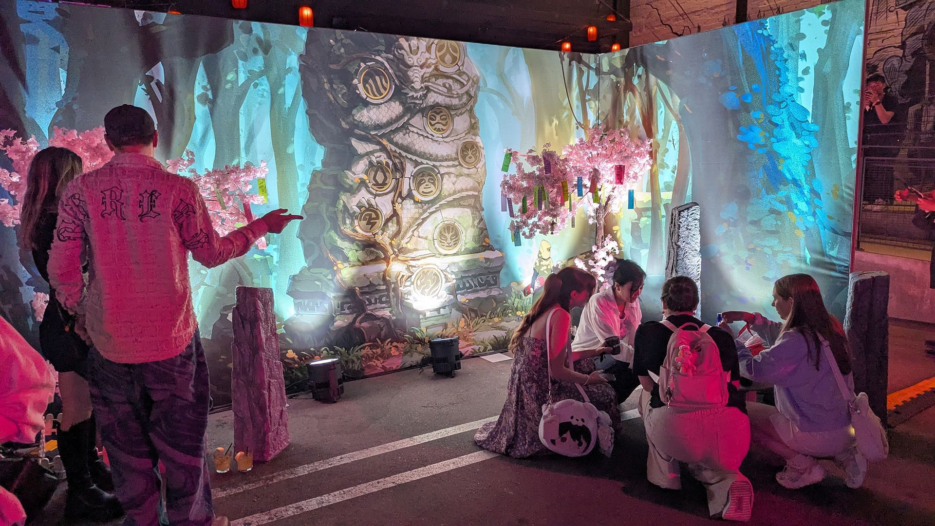 People interact with a colorful forest backdrop. They gather around a small, decorated structure.