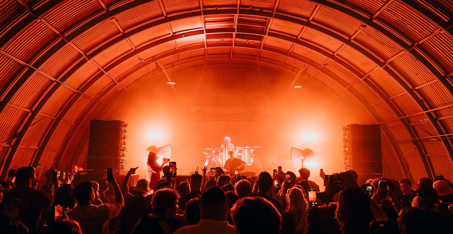 A crowd at a music concert in a red-lit arched building, people with arms raised, DJ on stage.