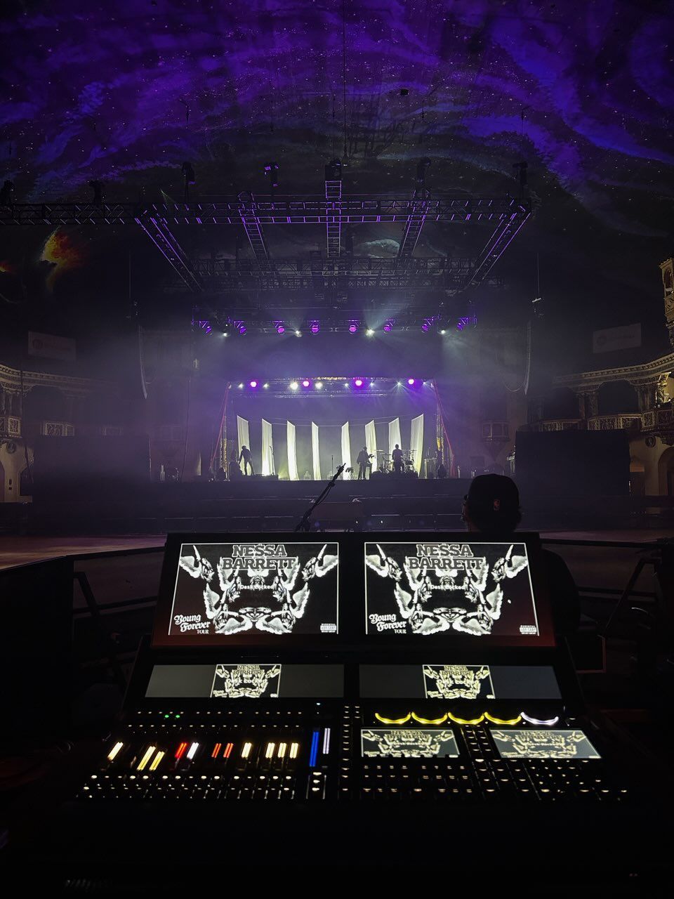 Control board view of a concert stage, bathed in purple and white lights. A silhouette works the console.