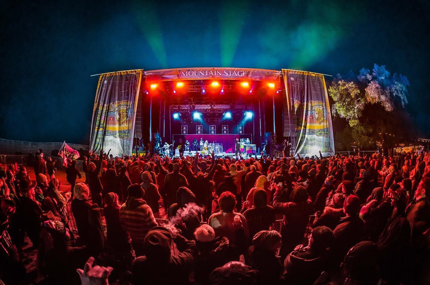 Concert crowd facing stage with bright lights. Band performing on stage at outdoor festival.