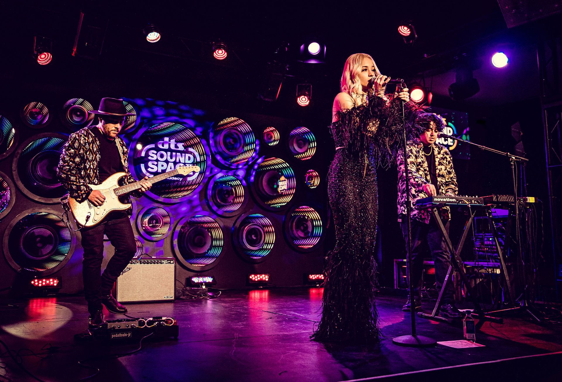 A band performs on stage with a singer in a sparkly dress, guitar, and keyboard players, colorful lights.