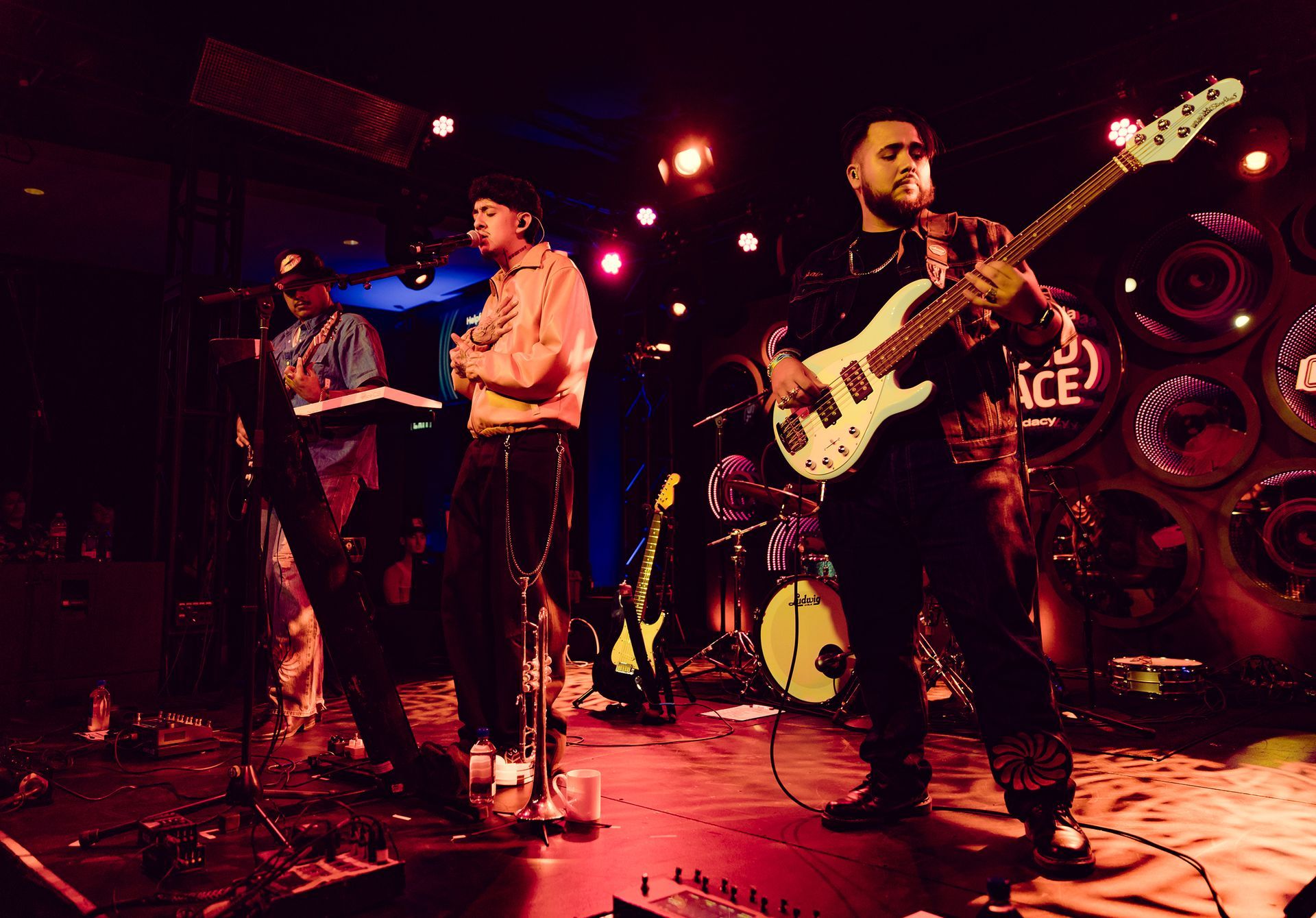 Band performing on stage, with singer, keyboardist, and bassist. Red and blue stage lighting.