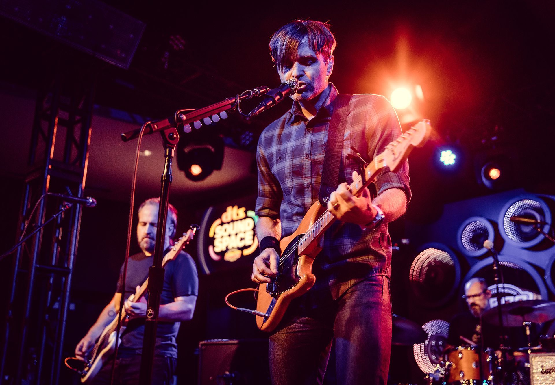 A band performing live on stage under bright stage lights. The lead singer plays guitar, other band members play bass and drums.