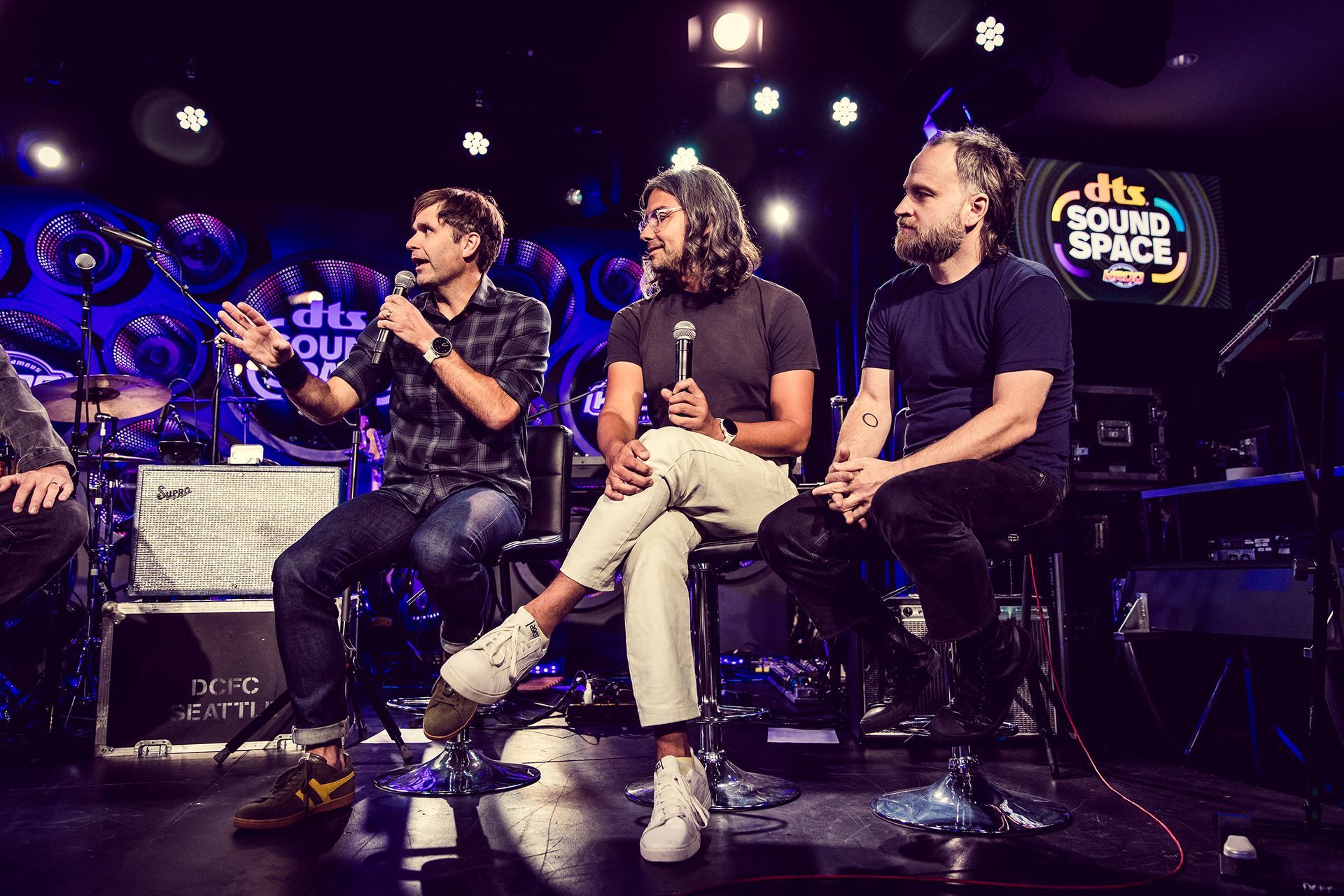 Three people on stools at a DTS Sound Space event. One man speaks, two listen. Stage setting with instruments.