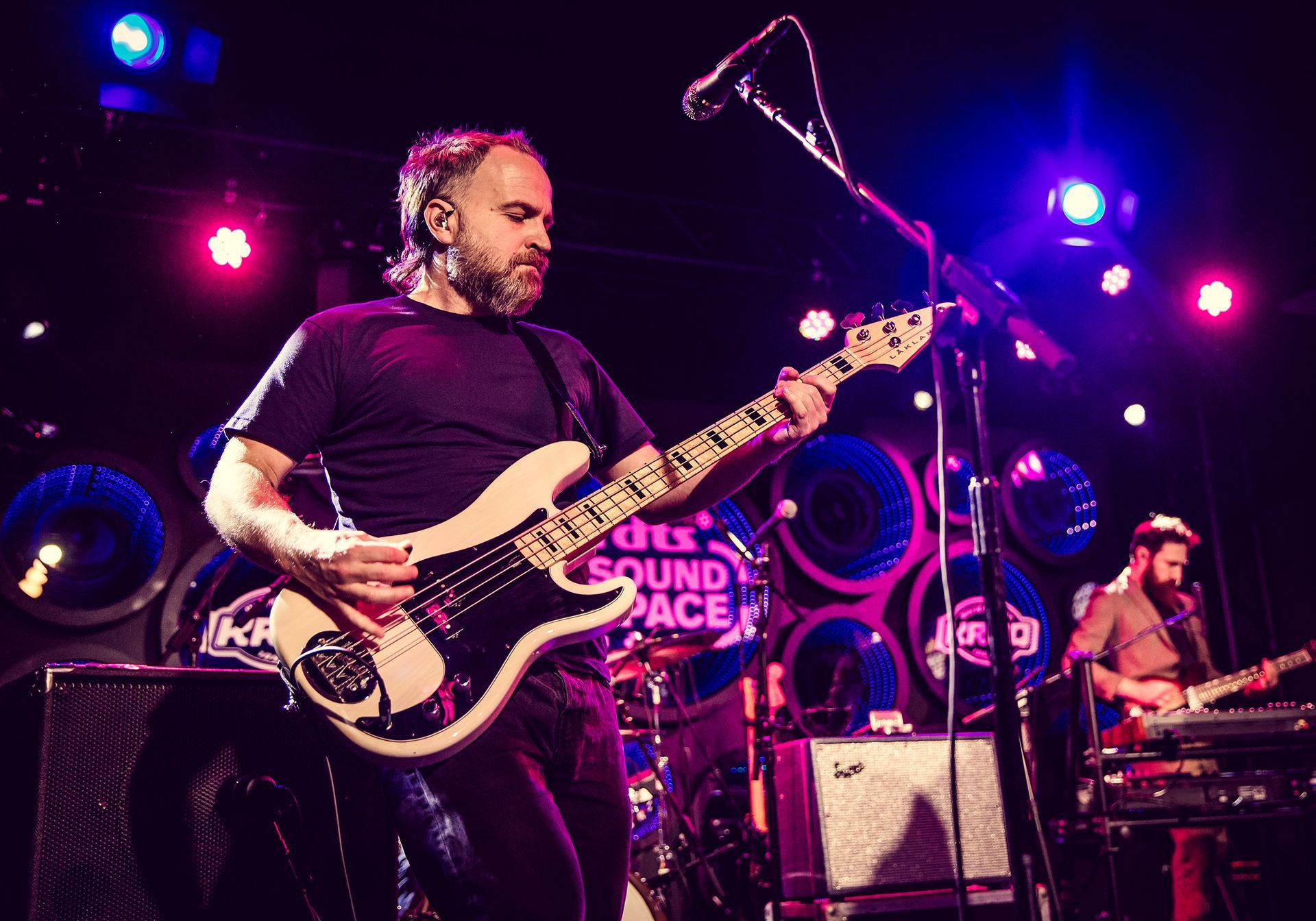A man plays a white bass guitar on stage, lit by blue and purple lights. Another musician is in the background.