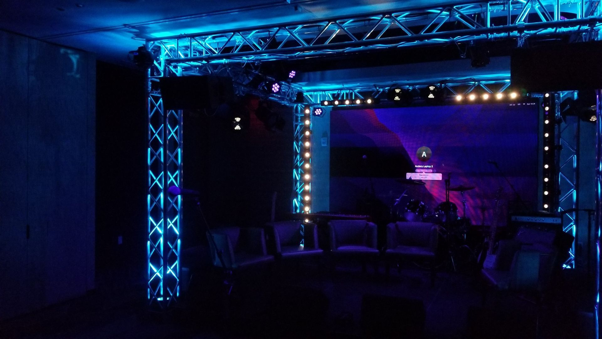 A dimly lit stage with blue lighting, truss system, and a large screen displaying a purple graphic.