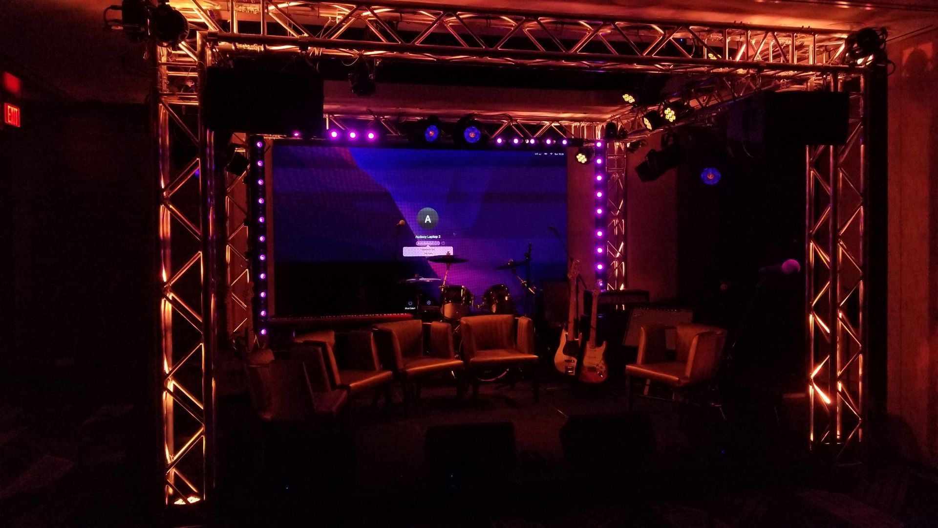 Stage set with lighting rig, screen backdrop, chairs, and guitars. Lit in orange and blue.