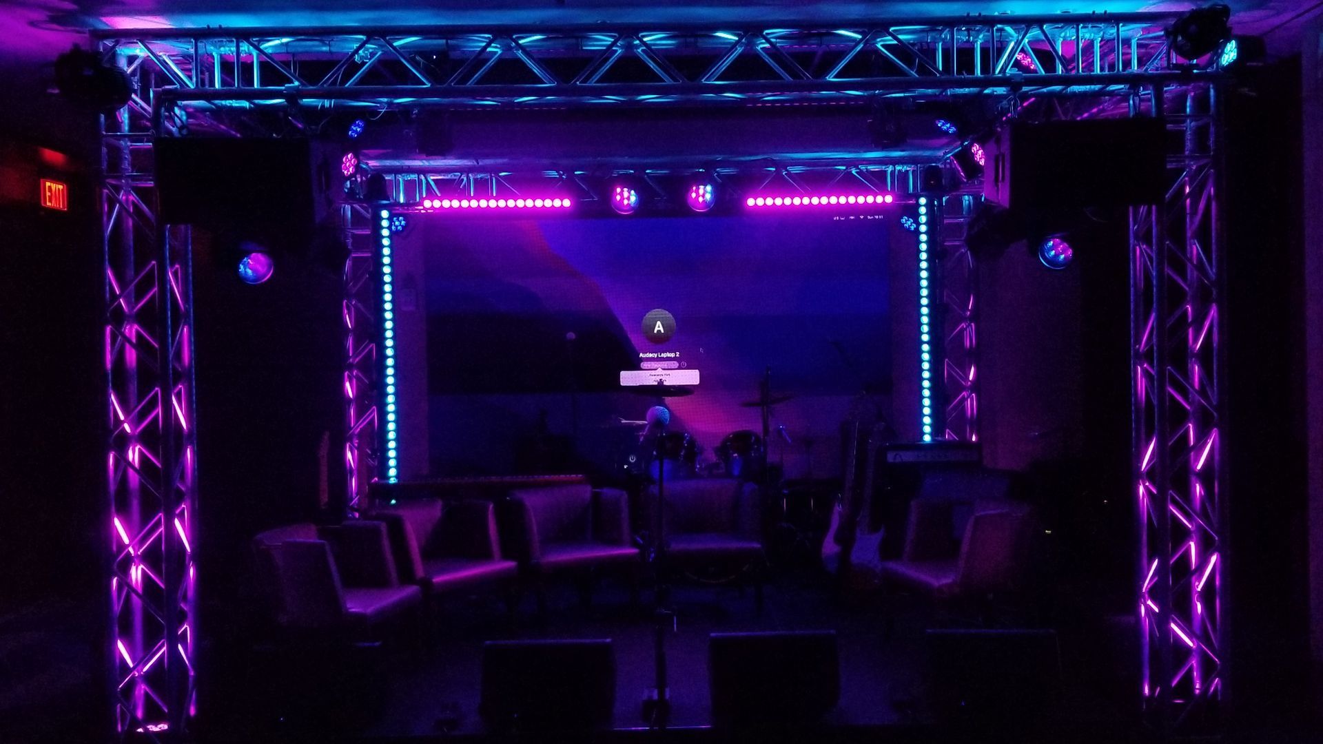 Lit-up stage setup with purple and blue lighting. Truss frame, speakers, and a screen in a dark room.