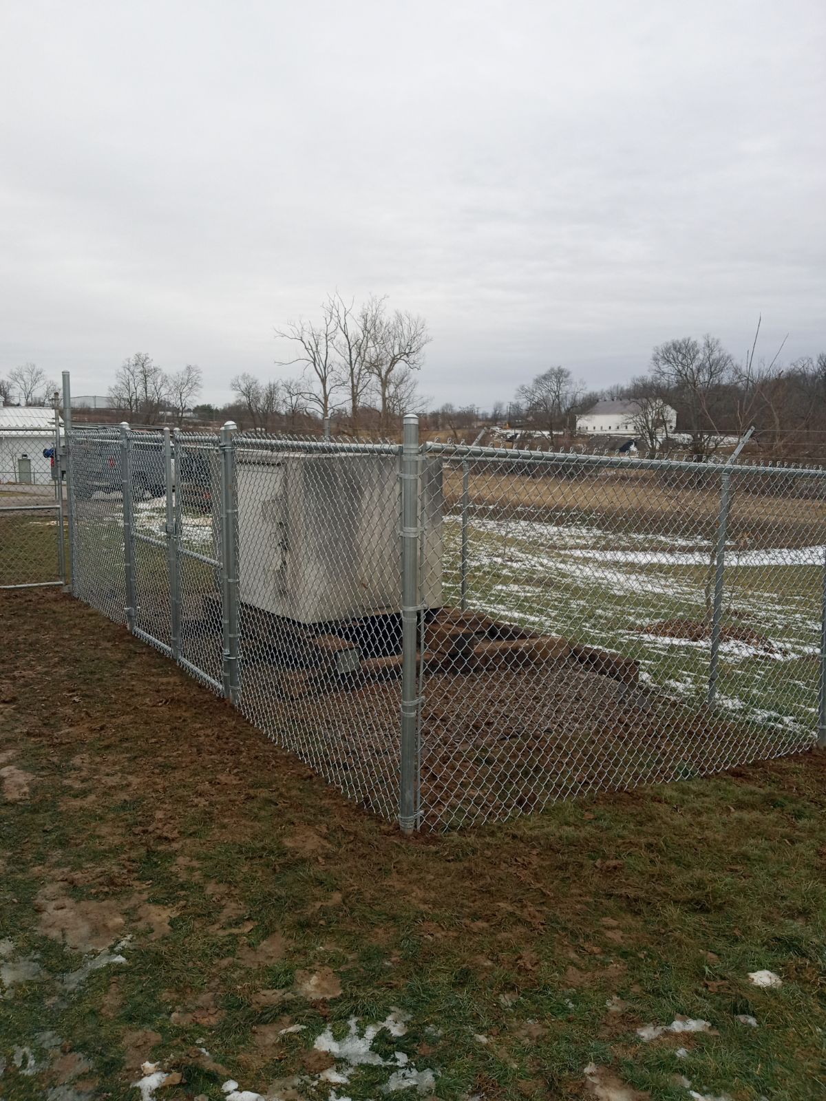 Chain-link fenced utility enclosure in a muddy field under a gray sky.