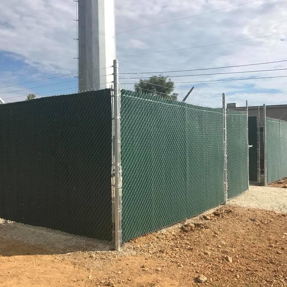 Green chain-link fence around a utility area on a dirt lot under a cloudy sky