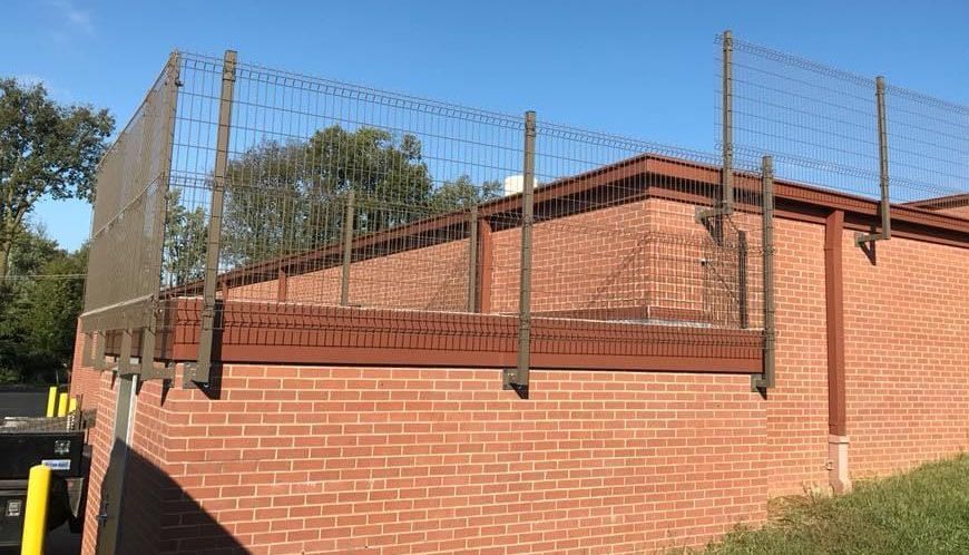 Brick building with tall metal mesh fencing along the roofline under a clear blue sky