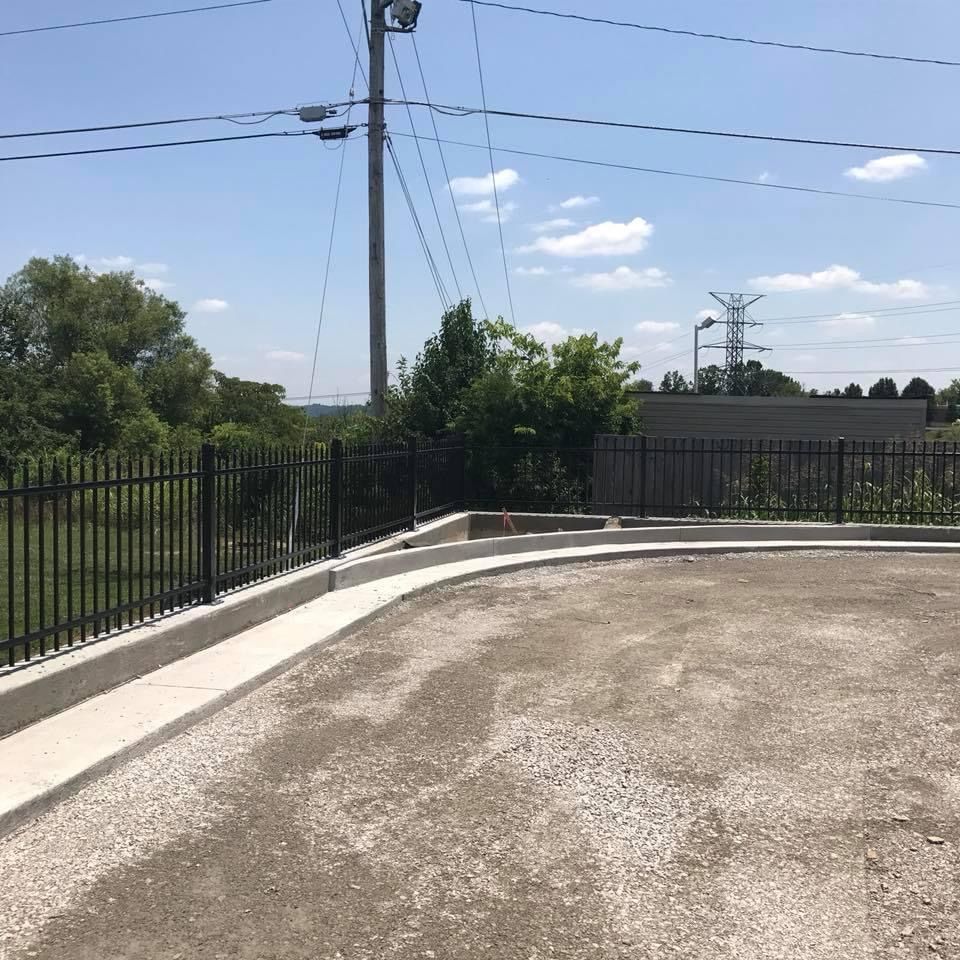 Curved paved driveway beside black metal fence under blue sky with utility poles and trees
