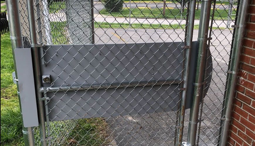 Chain-link fence gate with gray panels beside a brick building, viewed from the sidewalk.