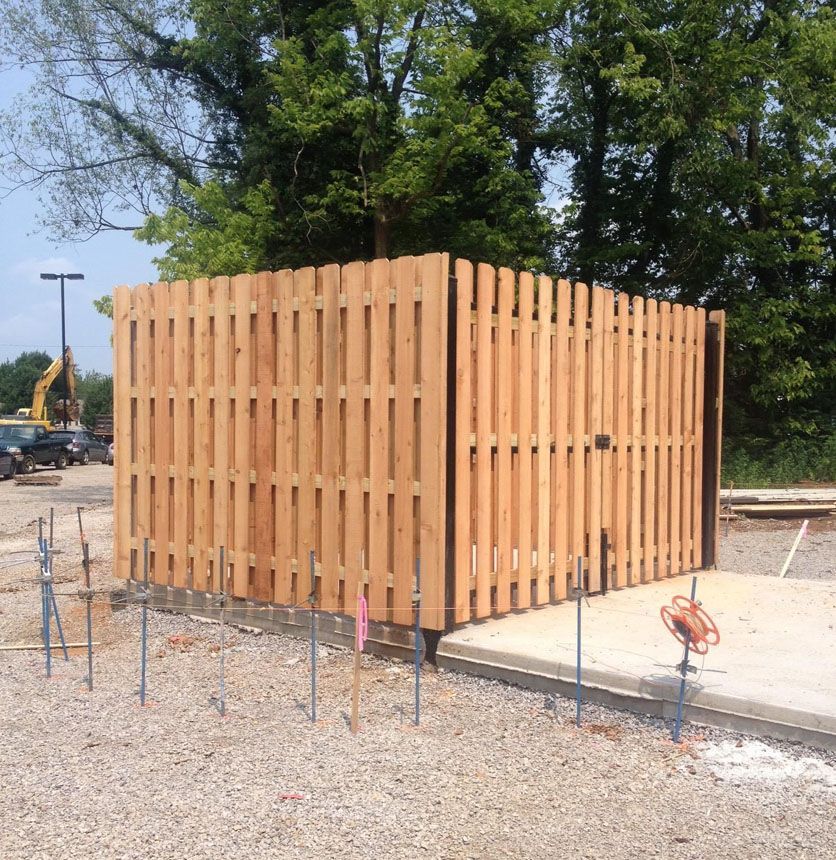 Wooden fenced utility enclosure on a gravel construction site, with trees in the background.
