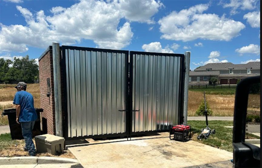 Two men working beside a large metal gate at a driveway under a partly cloudy sky
